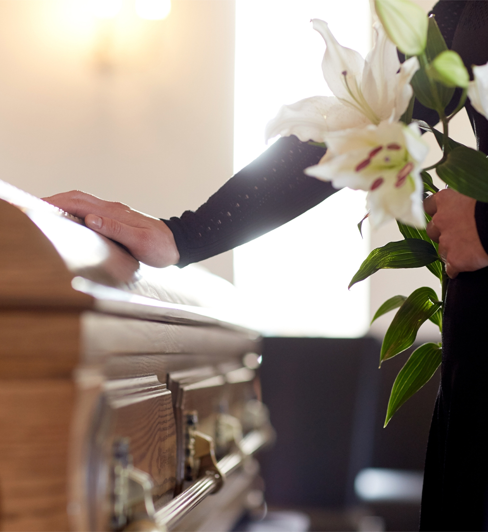 Person holds a lily with one hand while the other rests on a coffin