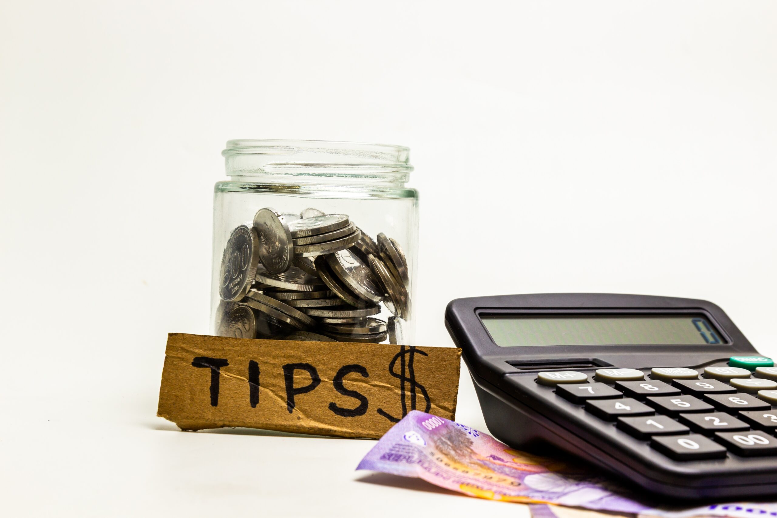 Jar of coins labeled “Tips” next to a calculator and cash, representing calculating reduced earnings for Niagara Falls service and hospitality workers